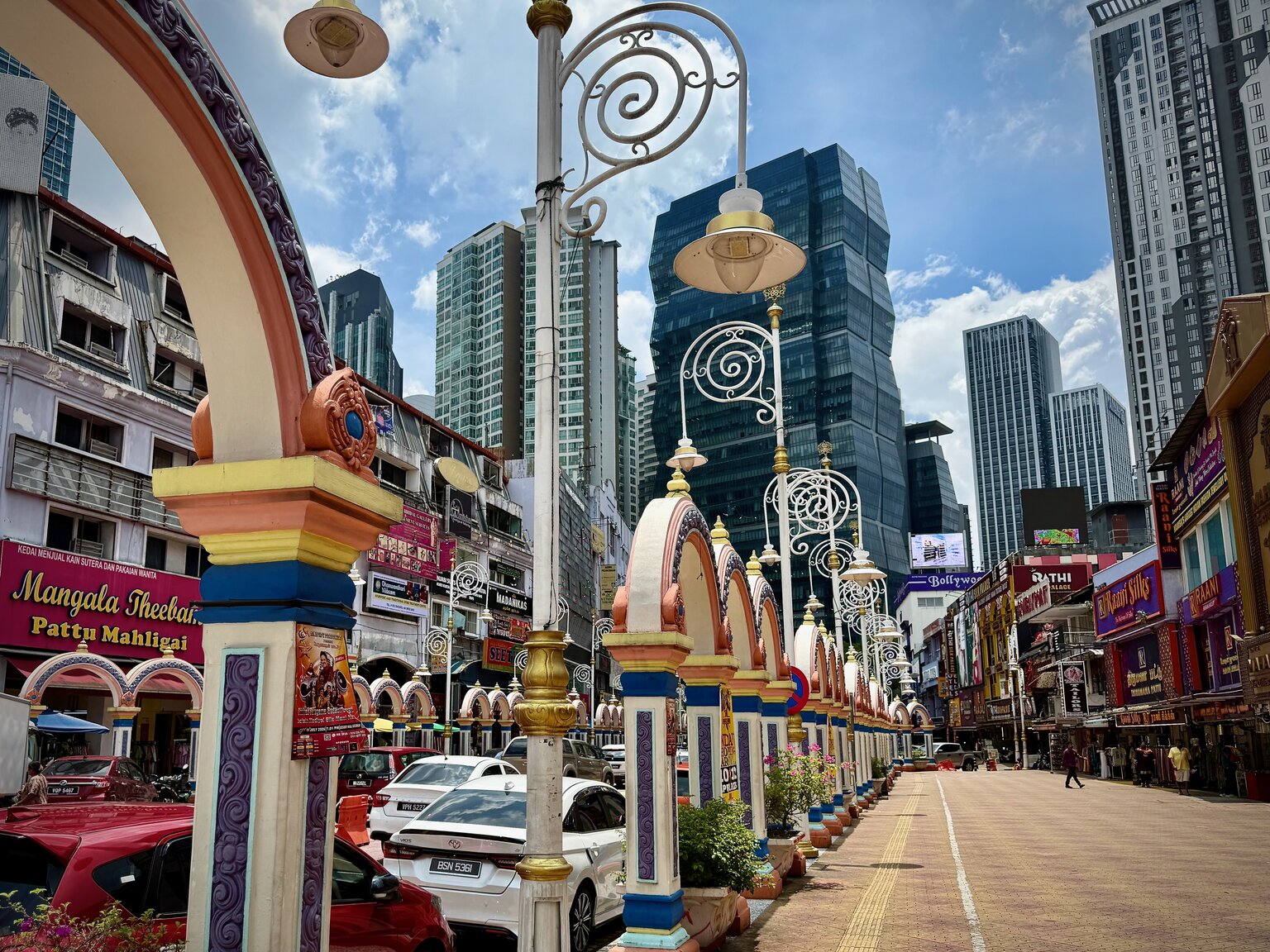 A street in Brickfields, Kuala Lumpur, lined with brightly painted ornate lampposts and decorated arches, with high-rises rising in the background
