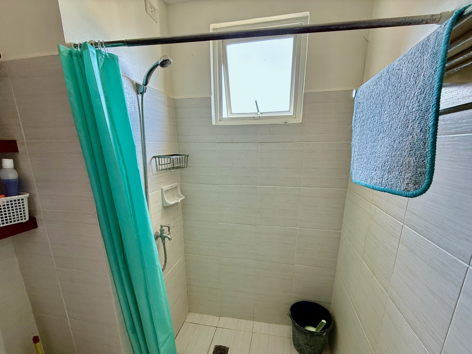 A simple tiled shower with a turquoise curtain, a towel hanging to dry, and a bucket on the floor.