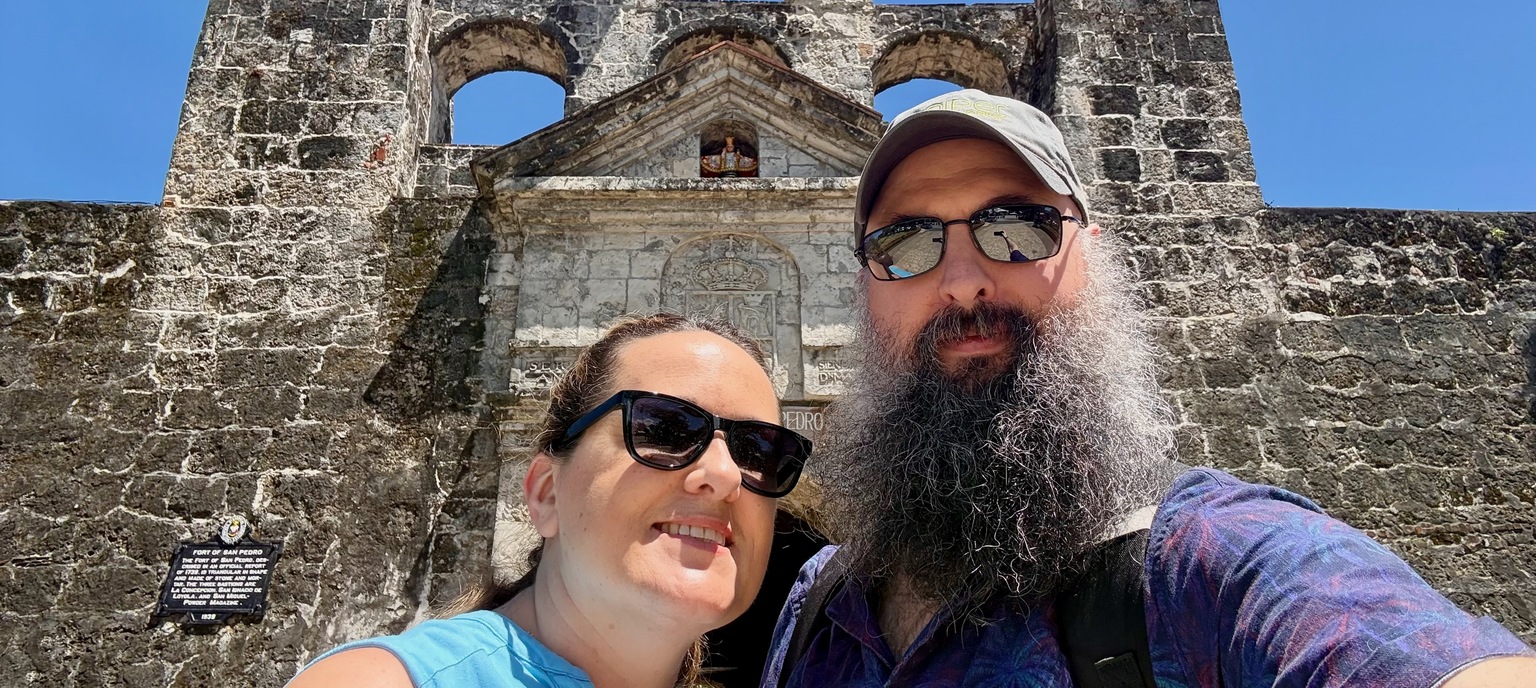 John and Marcia standing in front of Fort San Pedro in Cebu City under a bright blue sky.