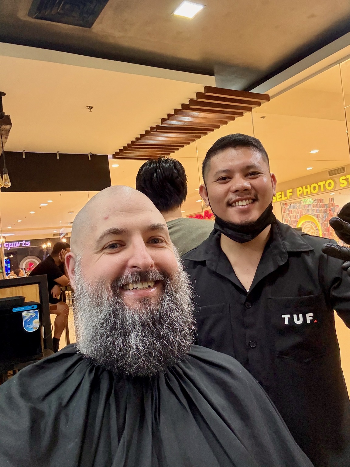 John smiling in a barber chair beside the barber after a haircut in a Cebu mall.