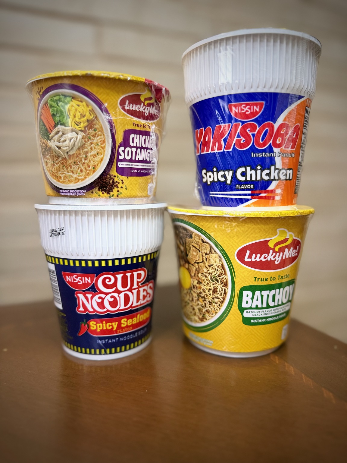 Four instant noodle cups stacked on a wooden table, including spicy chicken yakisoba, sotanghon, spicy seafood, and batchoy.