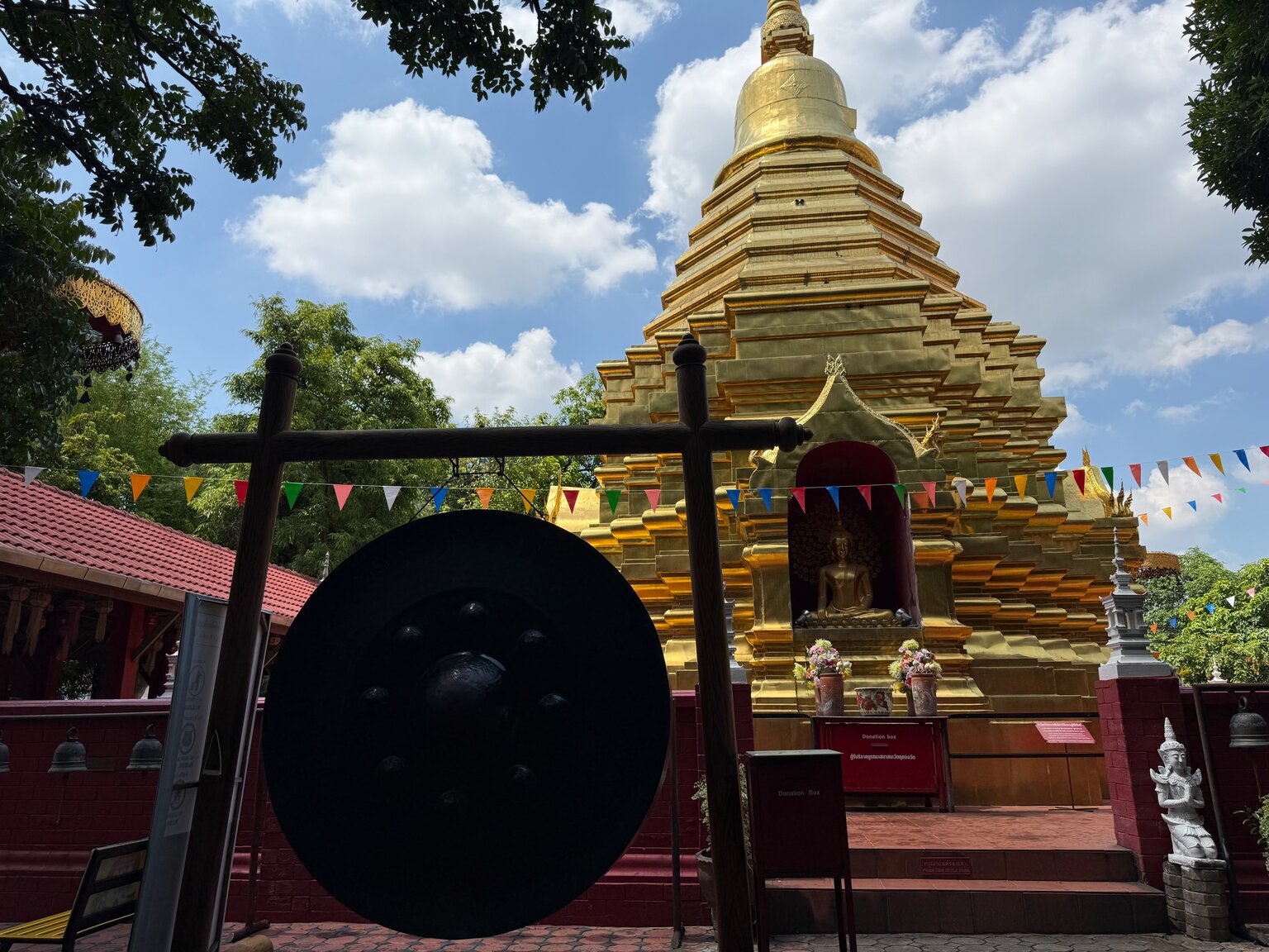 The golden stepped chedi at Wat Phan On under a bright blue sky, with strings of coloured prayer flags crossing the courtyard and a large black gong hanging in a wooden frame in the foreground.