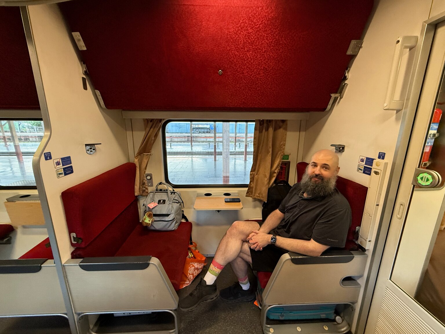 John sitting in a sleeper train compartment with red bench seats, a rucksack on the seat opposite and the station platform visible through the window