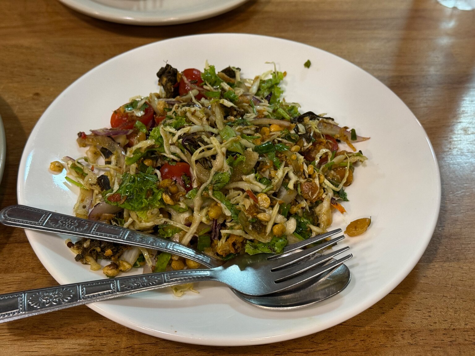 A plate of fermented tea leaf salad: shredded cabbage and tea leaves tossed with tomato, red onion, herbs, peanuts and crisp fried beans, a fork and spoon resting on the rim
