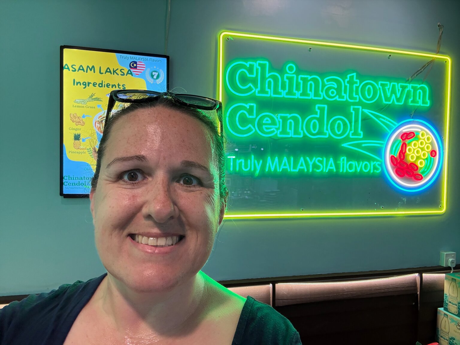 Marcia smiling in front of a bright green neon sign reading Chinatown Cendol, Truly Malaysia Flavors.
