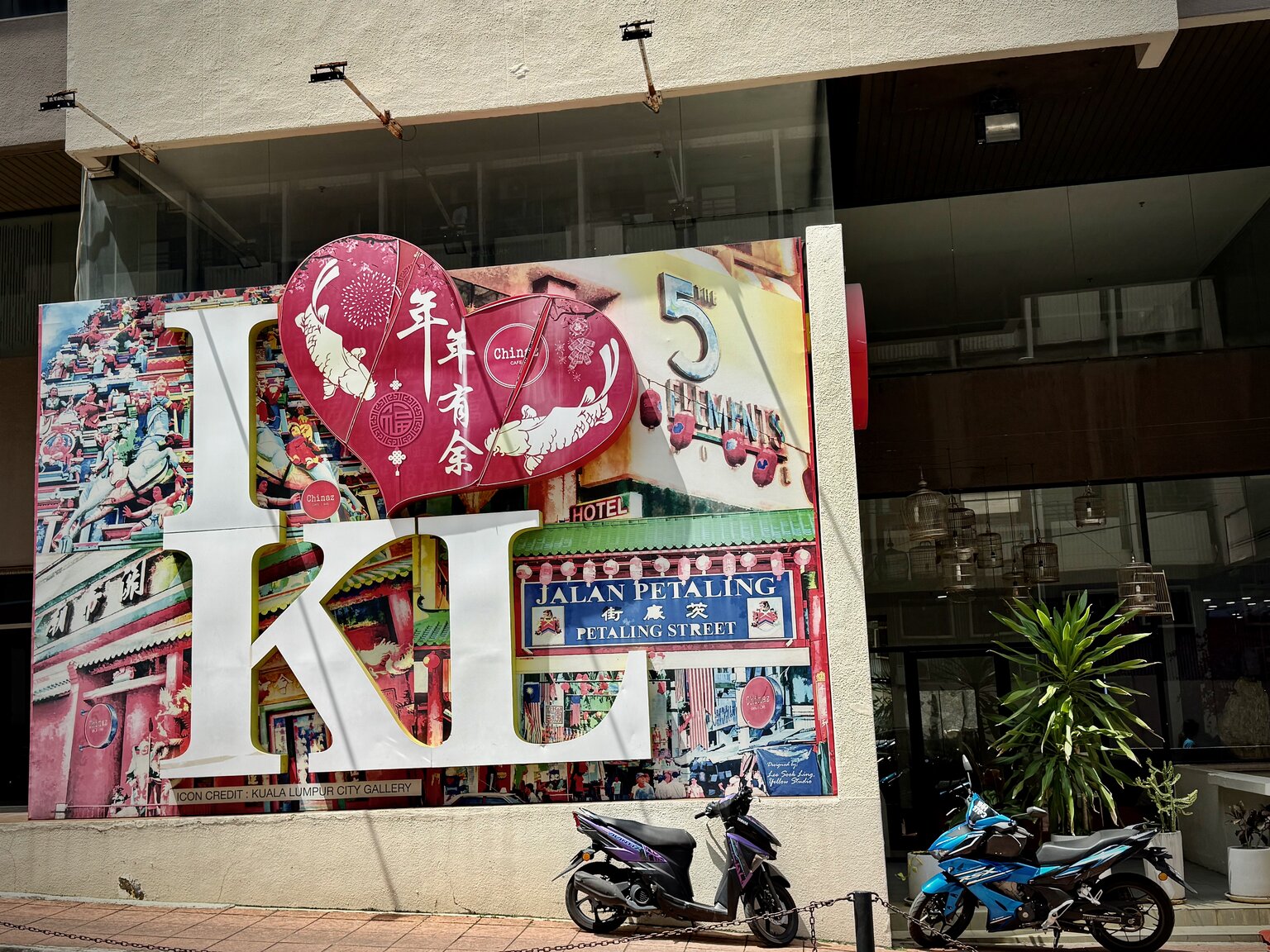 A large mural at the entrance to Petaling Street in Kuala Lumpur reading 'I love KL' with 'Jalan Petaling' painted along one side, and motorcycles parked in front of it