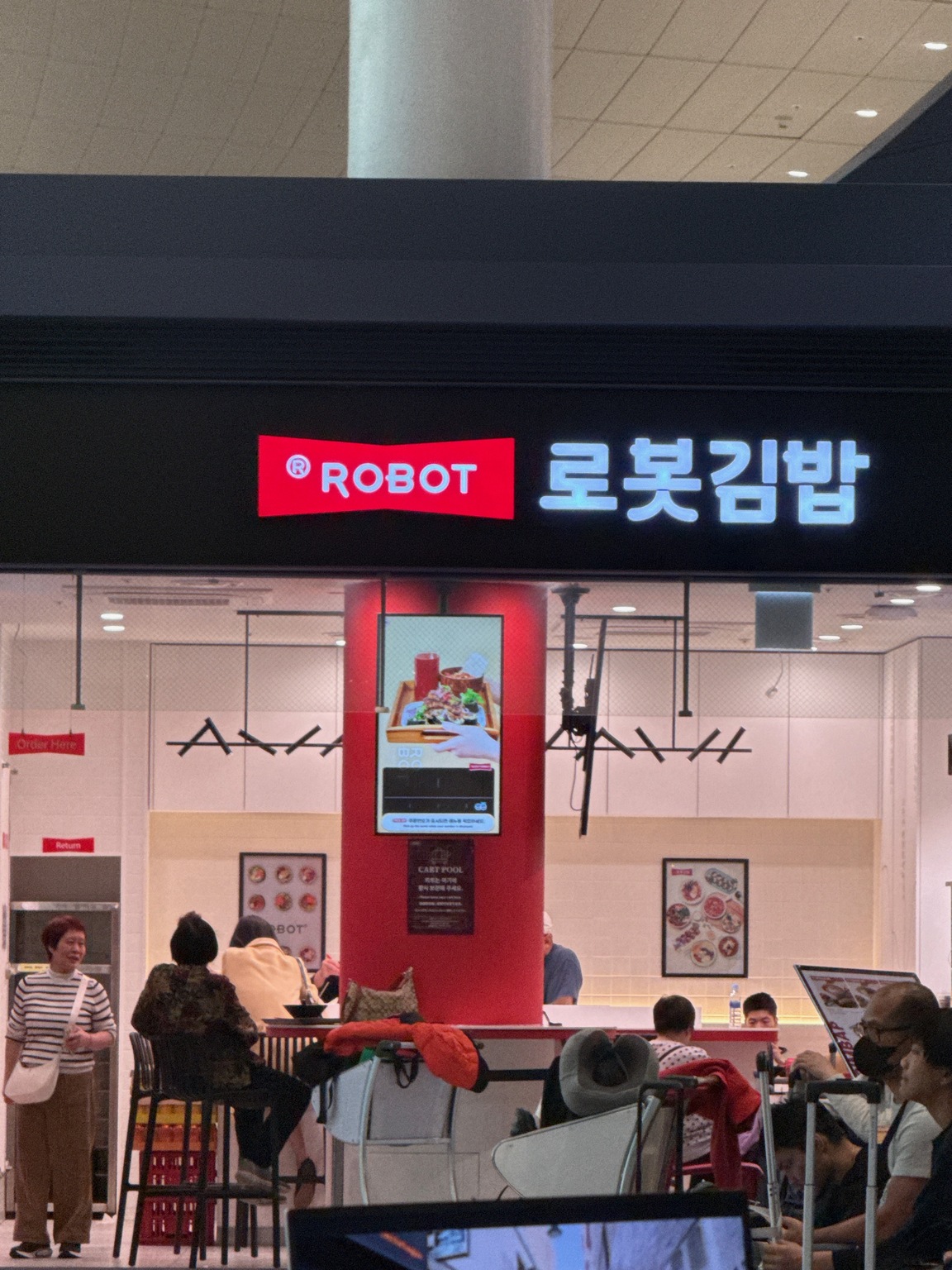 Robot gimbap restaurant in Incheon airport, with its illuminated sign, ordering screen, and travellers sitting nearby.