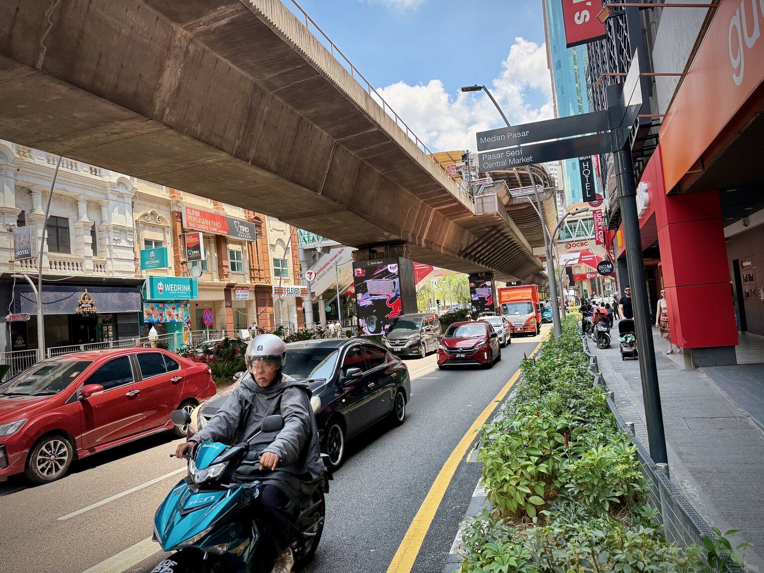 A busy Kuala Lumpur street running beneath an elevated LRT track, with motorcycles and cars in traffic below and shopfronts lining the road