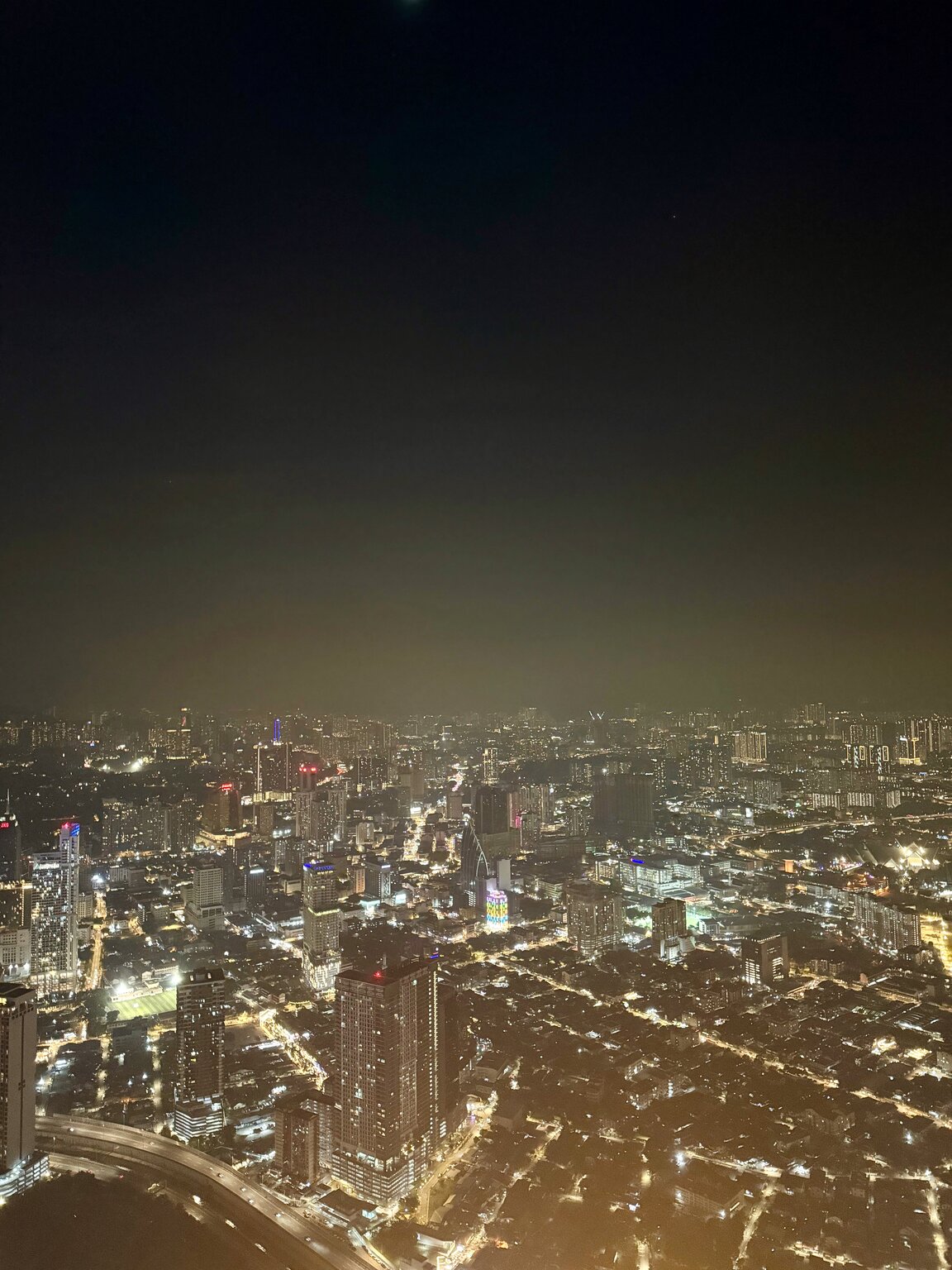 An aerial night view of Kuala Lumpur, the city lit up below with high-rises in the distance