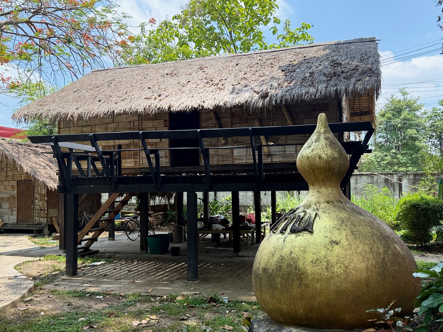A traditional Lanna house raised on wooden stilts, with a thatched roof and woven bamboo walls, and a large gourd-shaped clay jar standing in the foreground