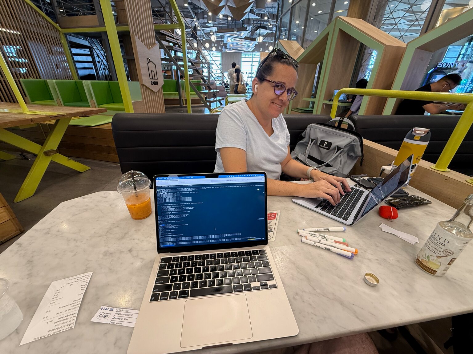 Marcia working at a laptop on a marble table in a coworking space, an iced drink beside her and the green and glass interior of the mall behind