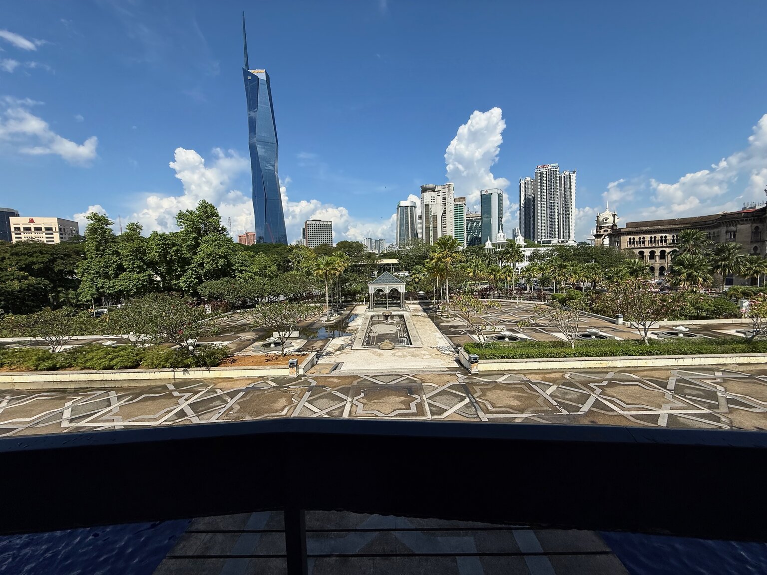 View from the National Mosque in Kuala Lumpur, looking out across a star-patterned tile floor and a stand of palm trees toward Merdeka 118 rising in the distance