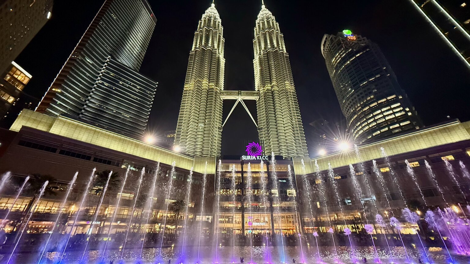 The illuminated Petronas Twin Towers at night rising above the lit KLCC fountain pool, with surrounding high-rises.