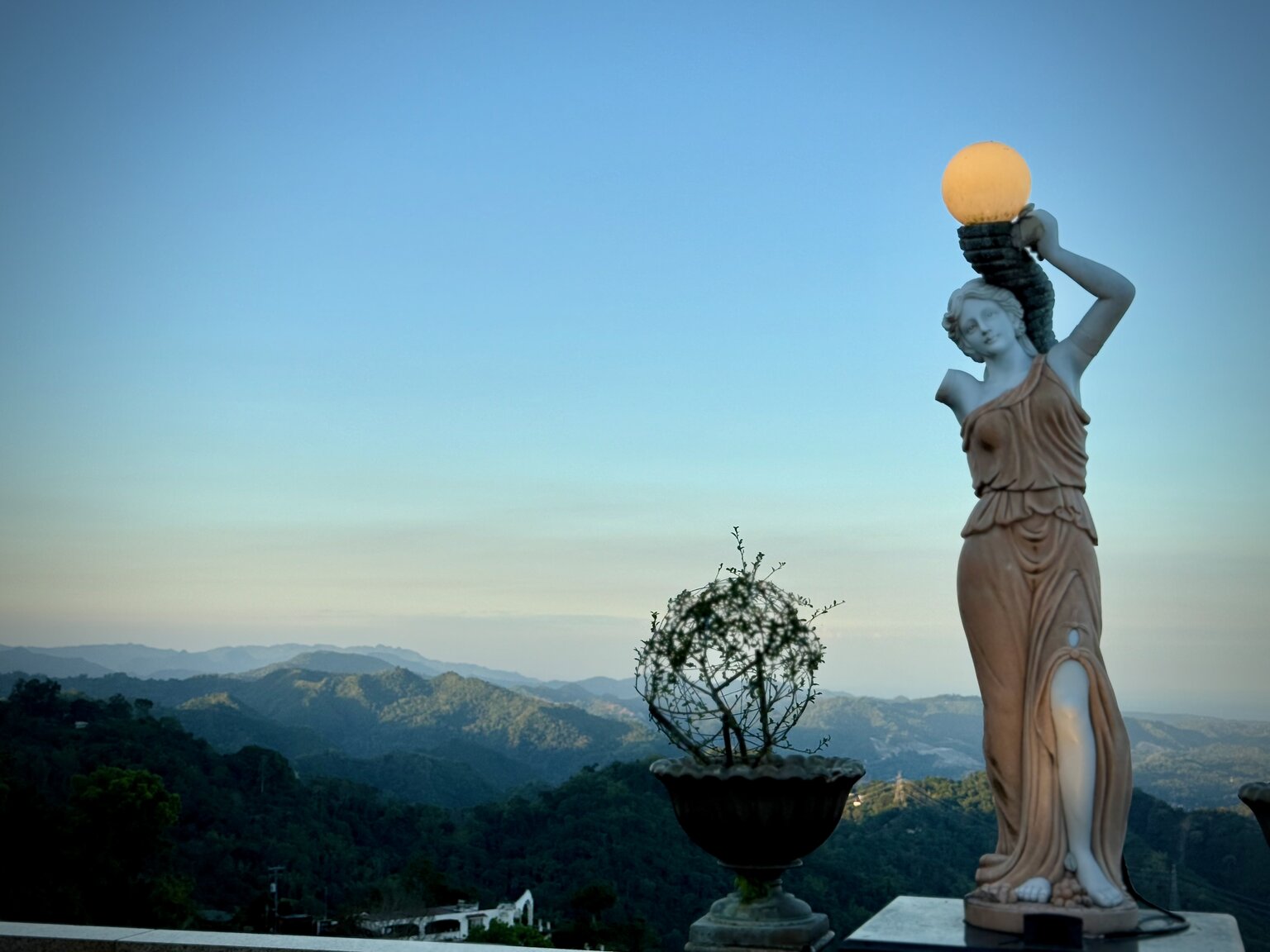 A classical marble statue of a woman holding a glowing orb lamp, lit at dusk with misty forested hills of Cebu spreading out behind her.