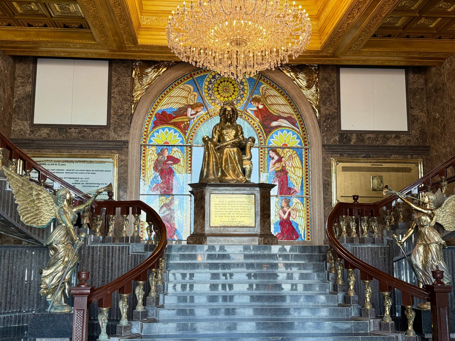 The grand entrance hall of the Temple of Leah: twin curved staircases flanked by golden angel statues, a crystal chandelier overhead, and a statue of Leah beneath a stained-glass window.